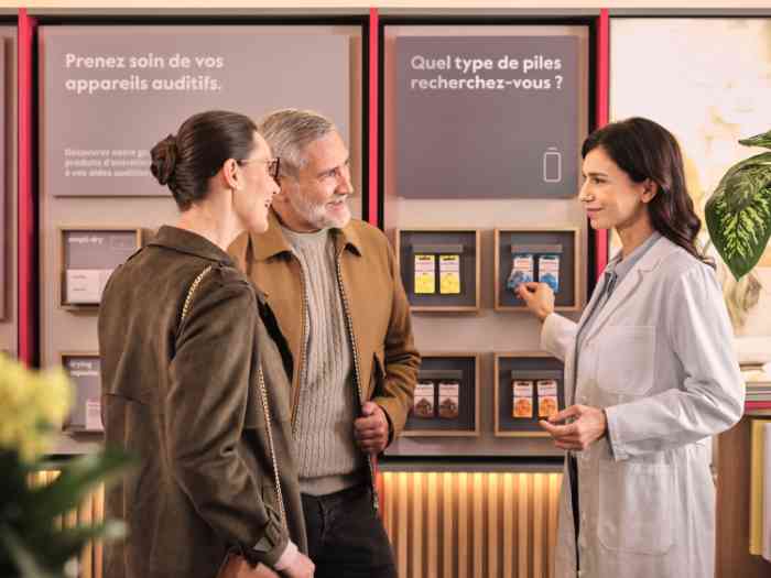 a couple and an audiologist in a lab coat talking in front of a panel displaying hearing aid batteries, french store.