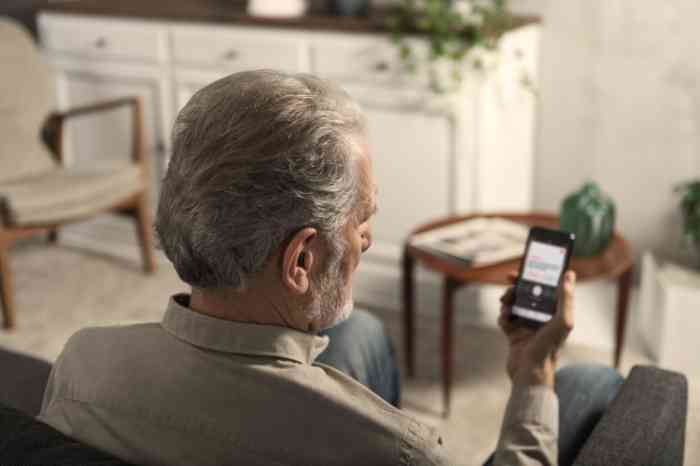 Homme âgé avec une aide auditive utilisant une application sur un smartphone sur un canapé.