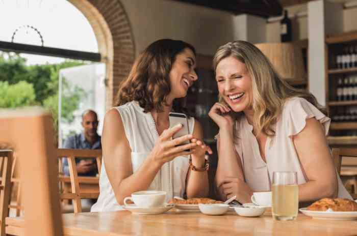 two women laughing and looking at a phone in a café.